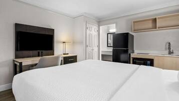 A hotel room with a large bed, a desk with a chair, a television, a refrigerator, a sink, and a mirror.