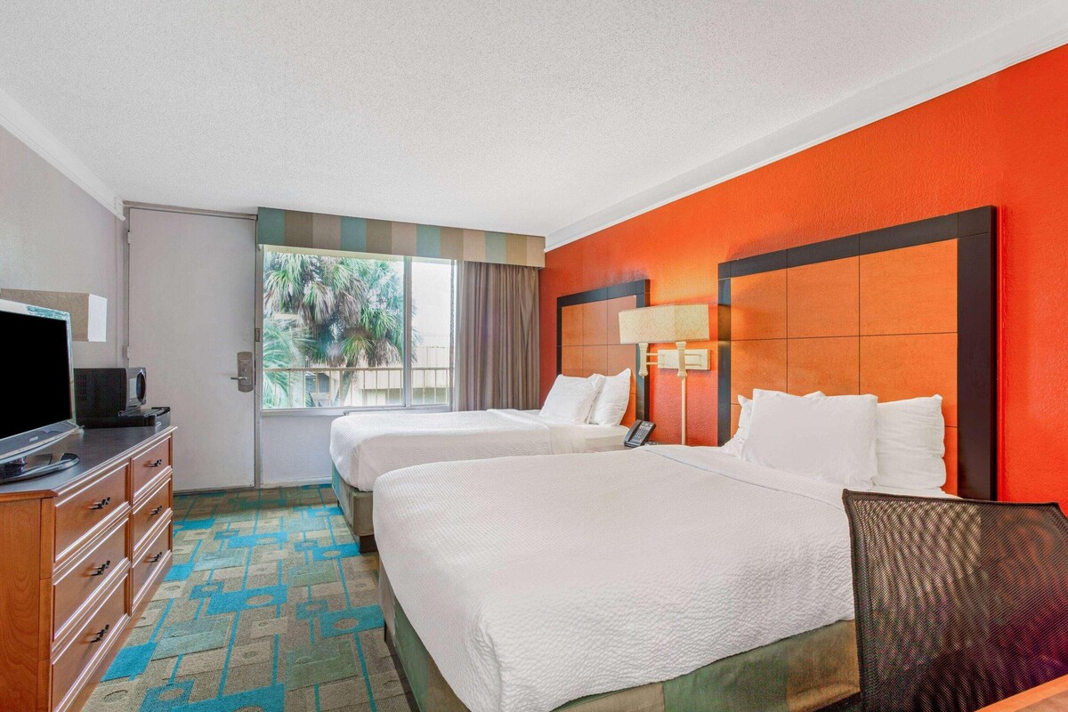 Habitación de hotel con dos camas, televisión, cómoda y ventana con vistas a palmeras.