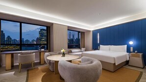 Una habitación de hotel moderna con una cama grande, un área de descanso y vistas a un paisaje urbano.
