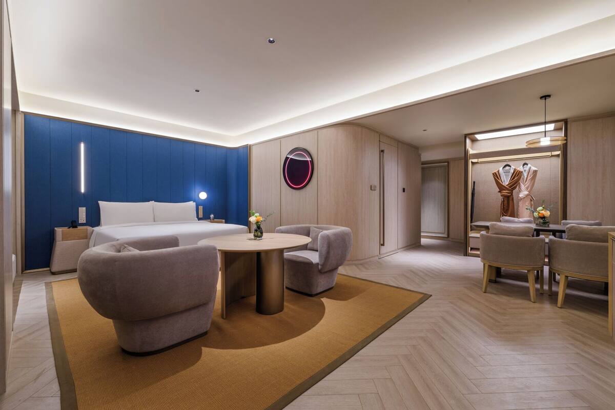 Una habitación de hotel moderna con una cama, dos sillones, una mesita y un perchero para bata y toallas.