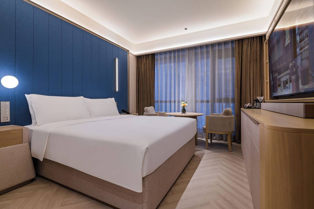 Habitación de hotel con una cama grande, un escritorio, una silla, un televisor y una ventana con cortinas.