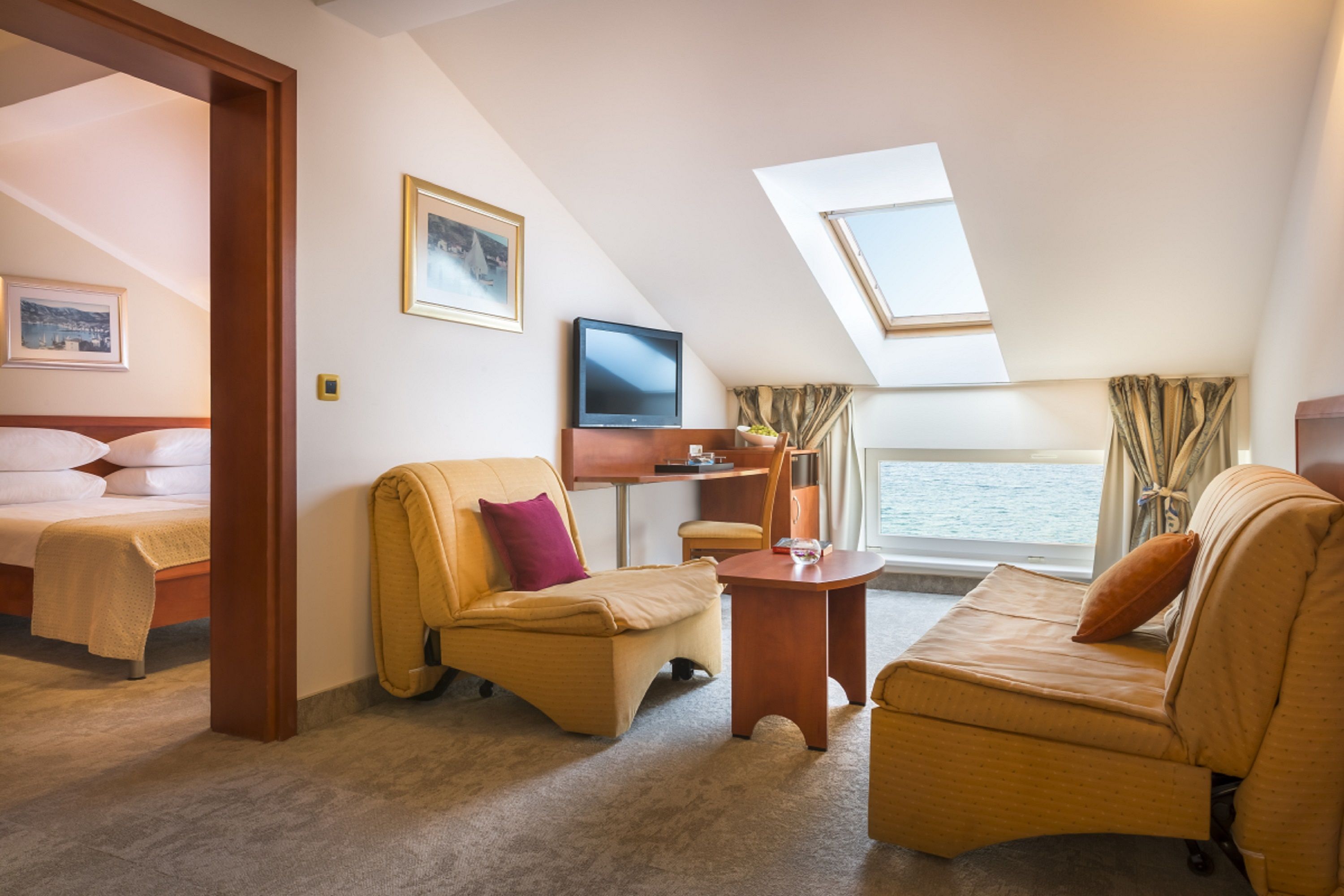 A room with a bed, two sofas, a television, a small table, and a window overlooking a body of water.
