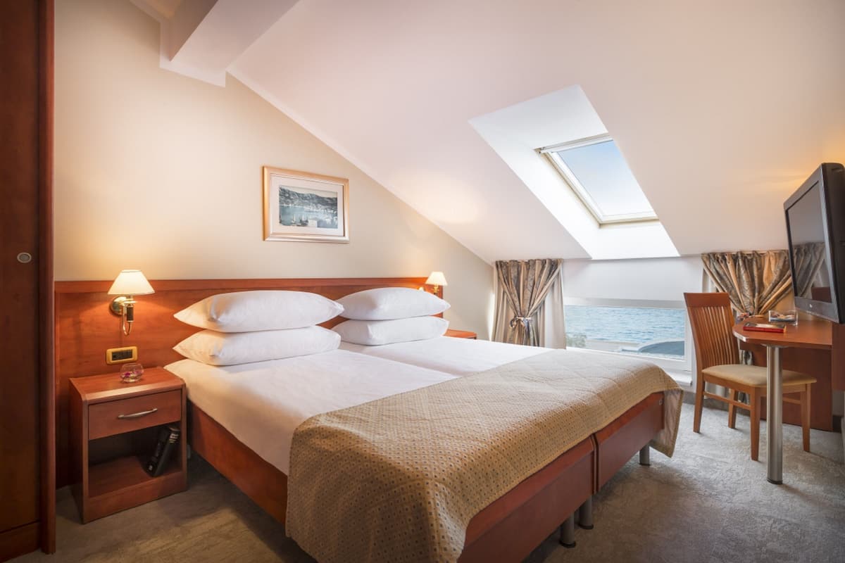 Habitación de hotel con cama, escritorio, silla, mesita de noche, lámpara, un cuadro enmarco y ventana con cortinas.