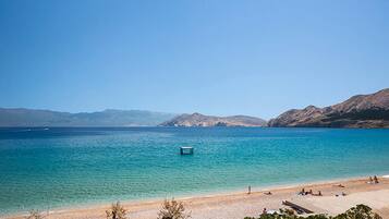 A beach with clear blue water, a few people, and mountains in the background.