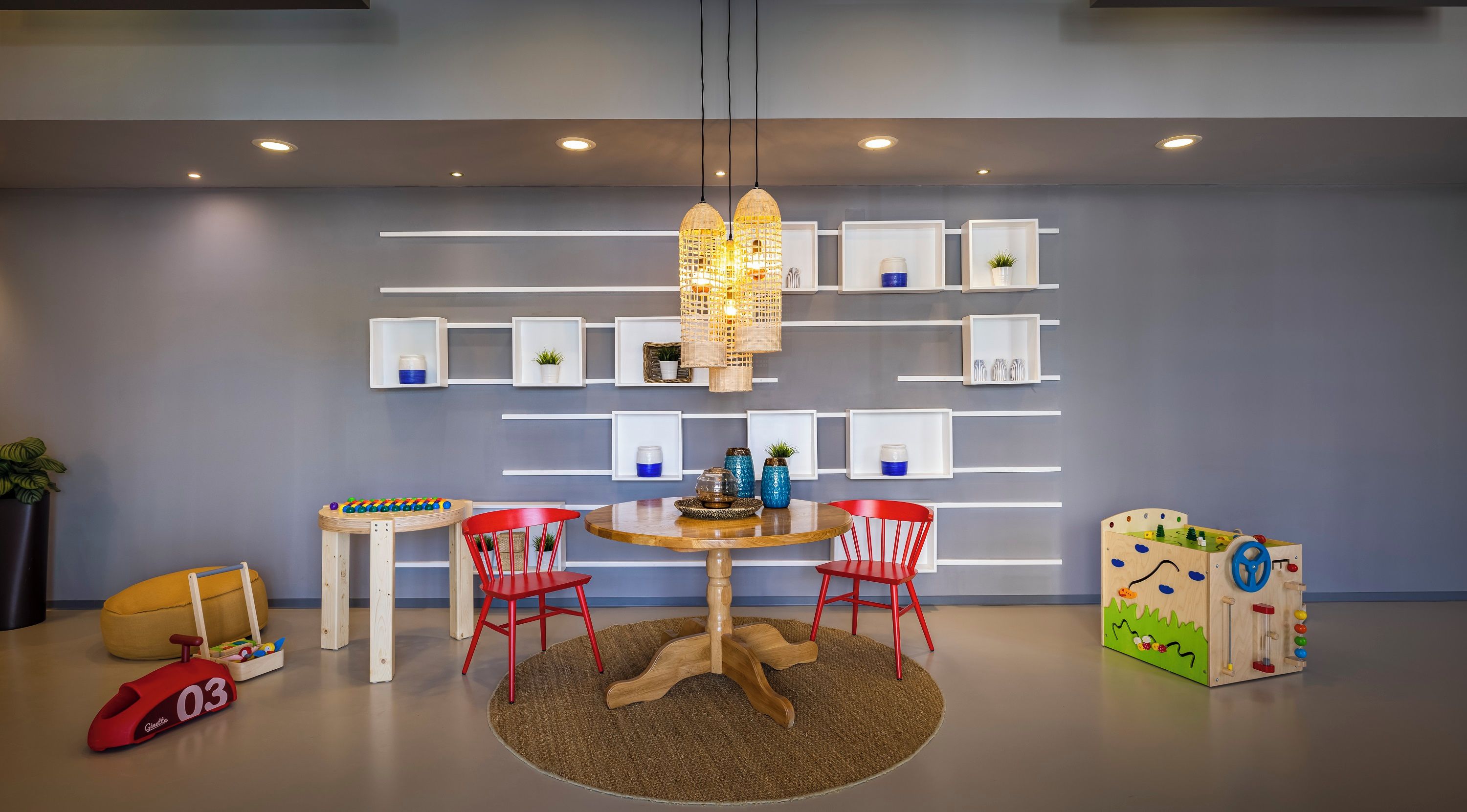 A modern children's play area with a round table, two red chairs, and a variety of toys and shelves against the wall.
