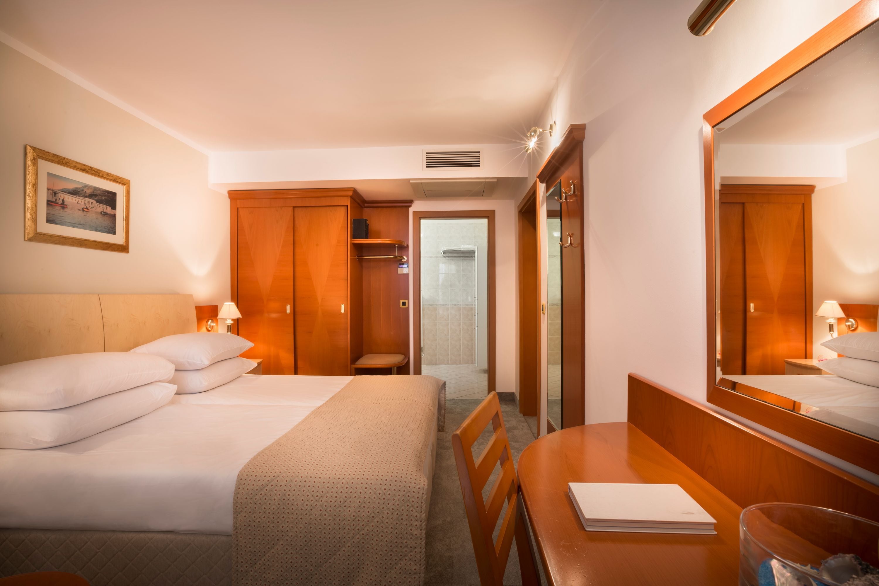 A hotel room with two beds, a desk, a chair, a mirror, a wardrobe, and a bathroom visible through an open door.