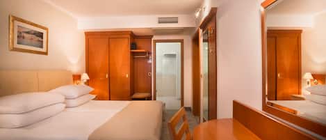 Habitación de hotel con dos camas, un escritorio, una silla, un espejo, un armario y un baño visible a través de una puerta abierta.