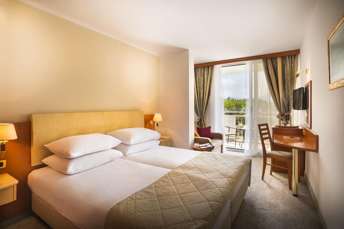 Habitación de hotel con cama, escritorio, silla, televisor y balcón con cortinas.
