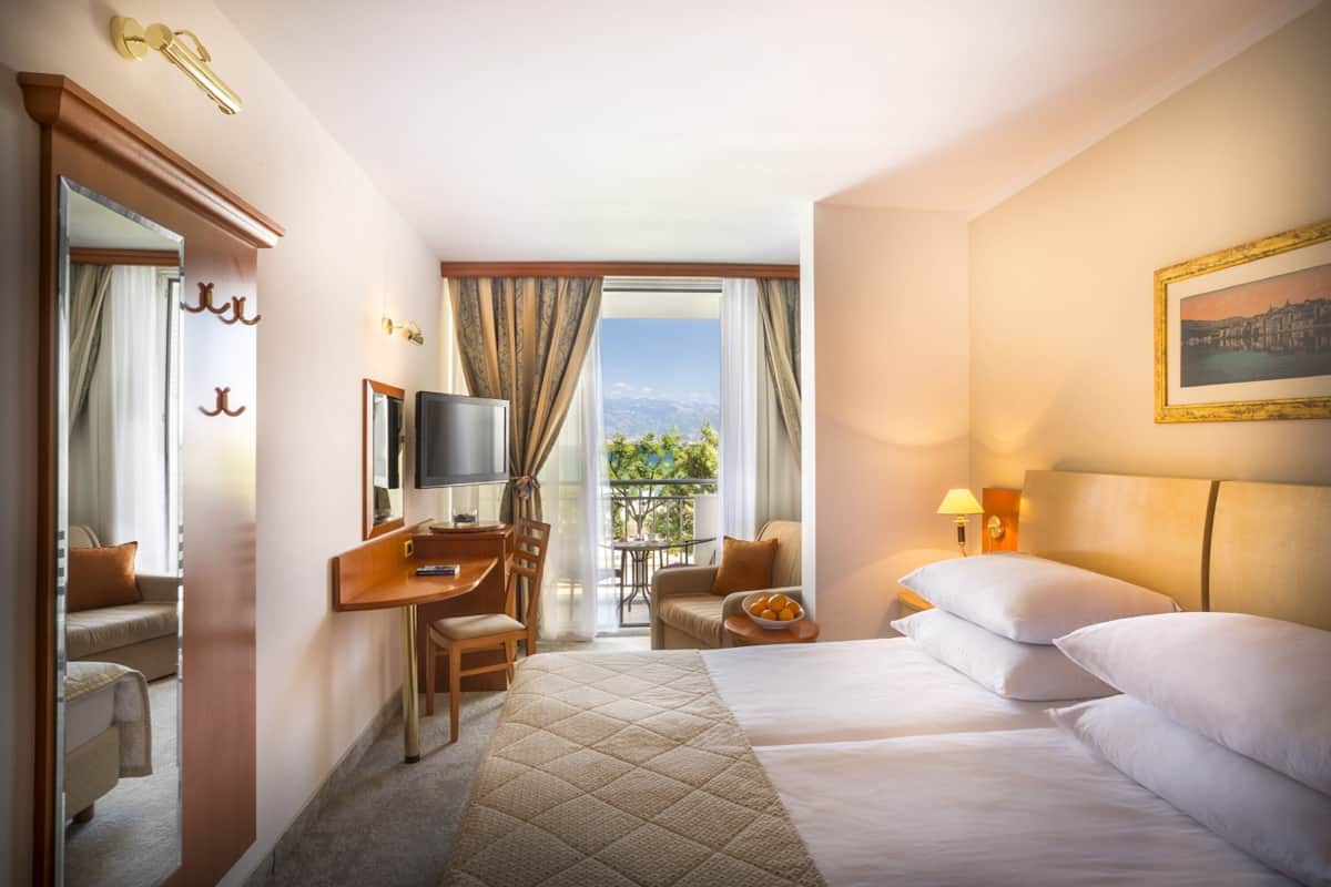 Habitación de hotel con cama, escritorio, silla, televisión, balcón con mesa y sillas, y un cuadro en la pared.