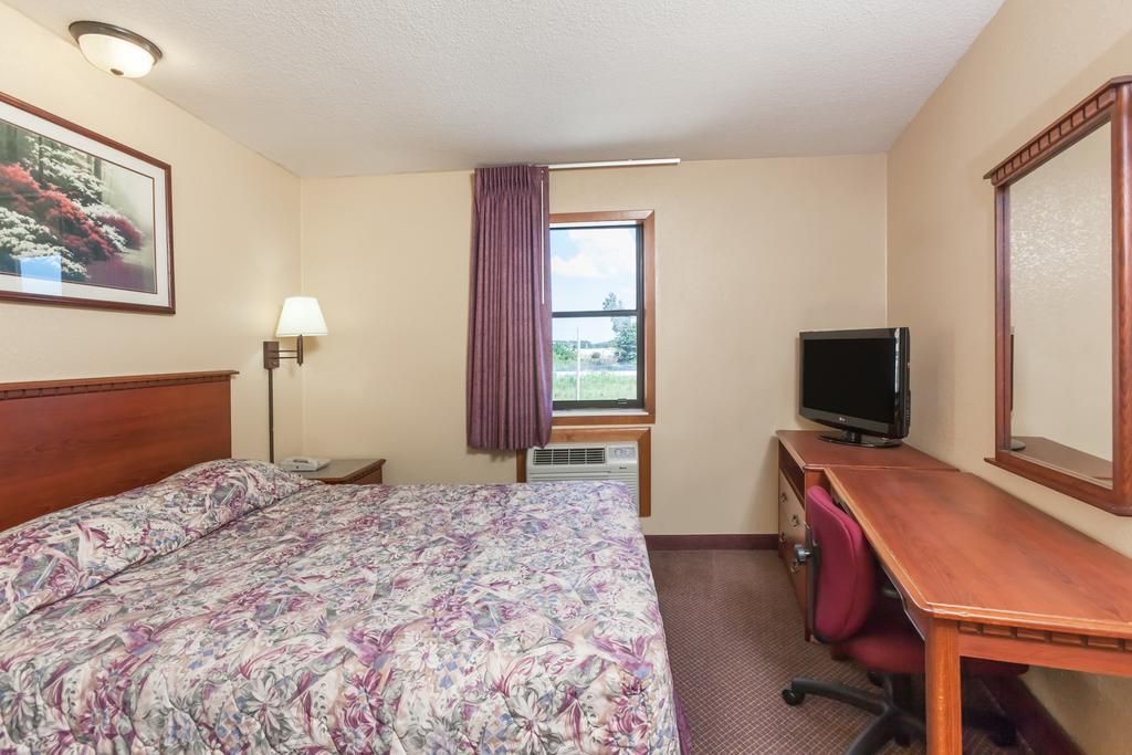 Habitación de hotel con cama, escritorio, televisor, lámpara y ventana con vistas a la vegetación.