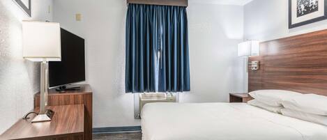 Una habitación de hotel con una cama, un escritorio con televisor, una lámpara de escritorio, una ventana con cortinas y un cuadro colgado en la pared.