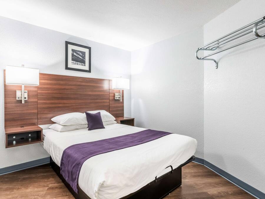 A hotel room with a wooden headboard, two bedside lamps, a bed with white and purple bedding, a small shelf, and a wall-mounted towel rack.