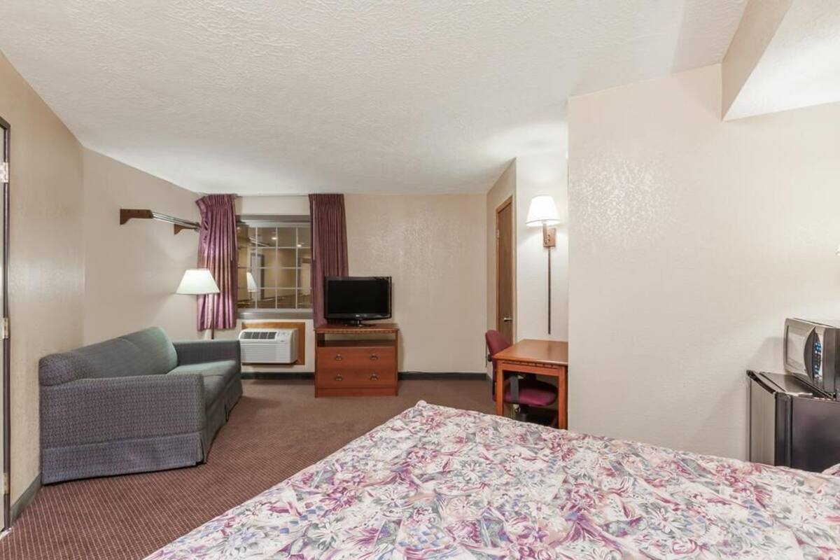 Quarto de hotel com cama, sofá, escrivaninha, televisão, abajur e janela com cortinas.
