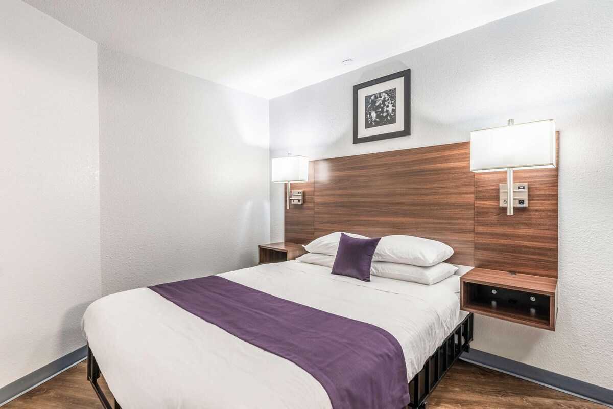A hotel room with a bed, a headboard, two bedside tables, a nightstand, a wall-mounted lamp, and a framed picture on the wall.