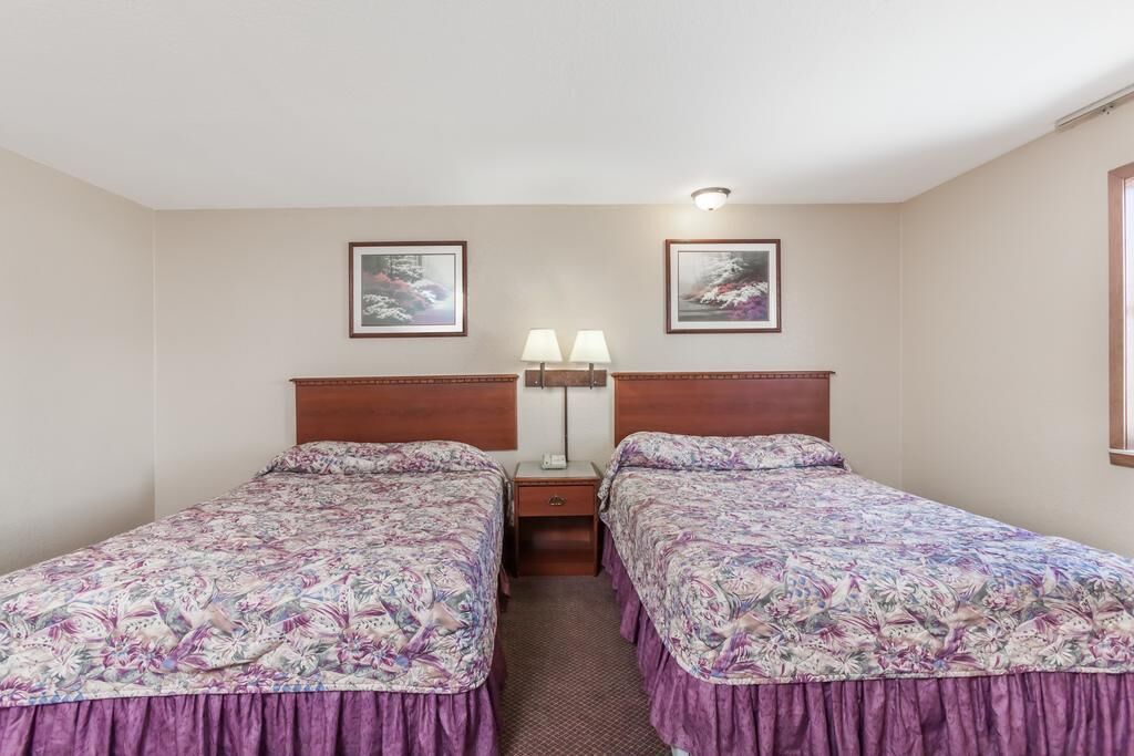 A room with two single beds, each with a floral bedspread and a wooden headboard. There is a nightstand with a lamp between the beds, and two framed pictures on the wall above.