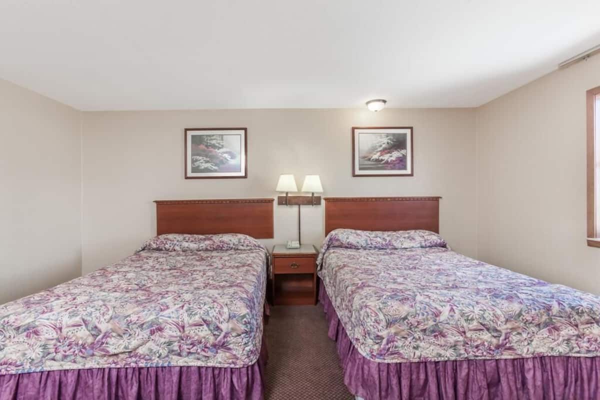 Quarto com duas camas de solteiro, cada uma com colcha florida e cabeceira de madeira. Há uma mesa de cabeceira com abajur entre as camas, e duas fotos emolduradas na parede acima.