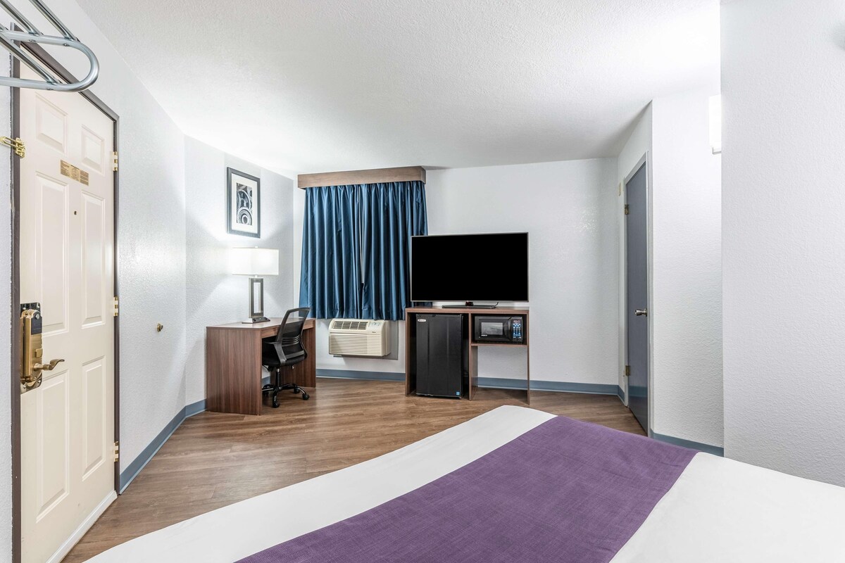 A hotel room with a bed, a desk with a computer, a TV, a mini-fridge, an air conditioner, and a door.