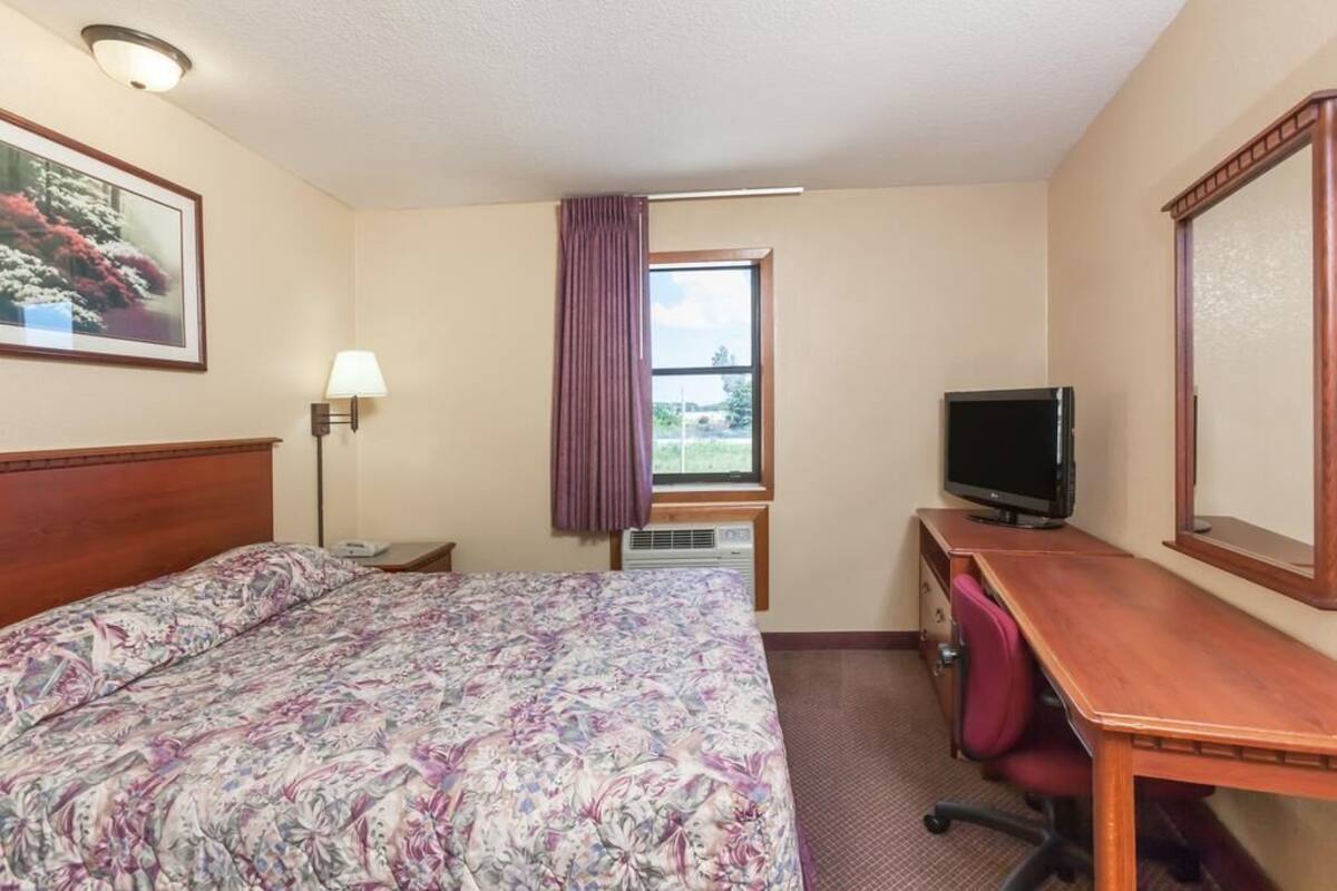 Quarto de hotel com cama, escrivaninha, televisão, luminária e janela com vista para vegetação.