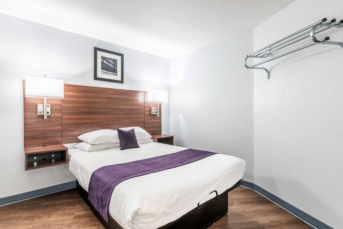 Quarto de hotel com cabeceira de madeira, duas luminárias de apoio, cama com roupa de cama branca e roxa, uma pequena prateleira e um porta-toalhas fixado na parede.
