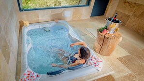A person relaxing in a hot tub with a view of a garden, a wooden stool with a bottle, and a glass of drink. - Hotel Le Domaine De Baulieu (Auch)