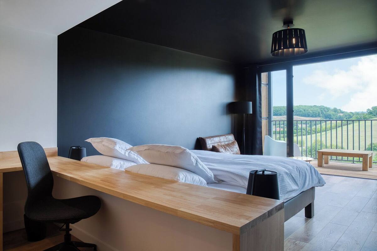 A modern bedroom with a large bed, a desk, and a chair. There is a balcony with a view of the countryside.