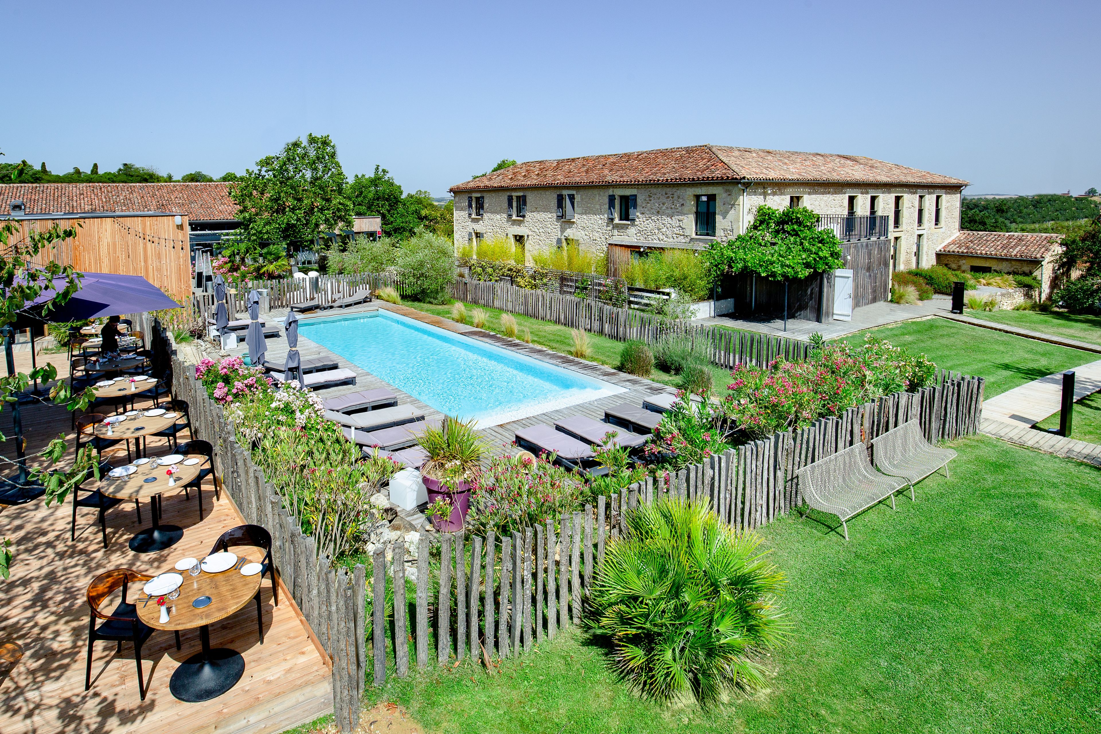 Un área junto a la piscina con una terraza de madera, sillas reclinables y un edificio de piedra al fondo.