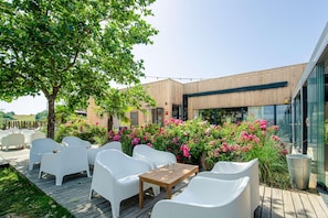 Un área de estar al aire libre con sillas blancas y una mesa de madera, rodeada de exuberante follaje y flores rosadas.