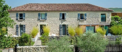 A two-story stone house with a tiled roof, a wooden deck, and a stone wall.
