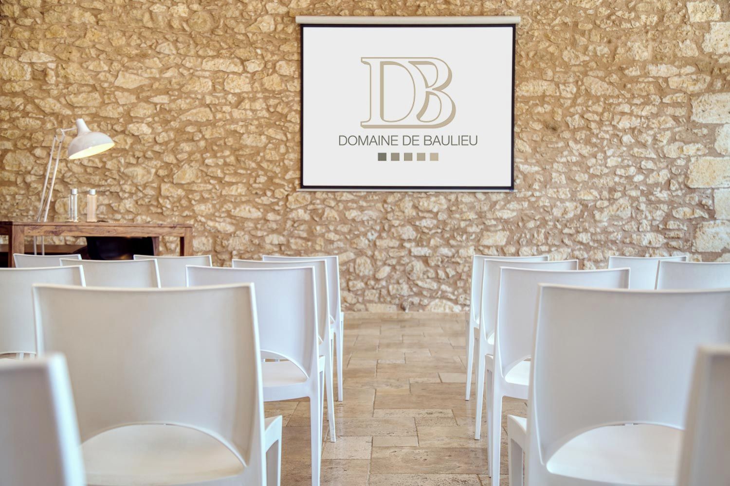 Interior de una pared de piedra con sillas blancas dispuestas para sentarse, un letrero enmarcado con el texto 'DB DOMAINE DE BAULIEU' y una mesa con una lámpara.