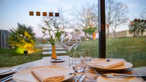 A dining table set with plates, cutlery, and wine glasses, with a view of a garden and trees outside. - Hotel Le Domaine De Baulieu (Auch)