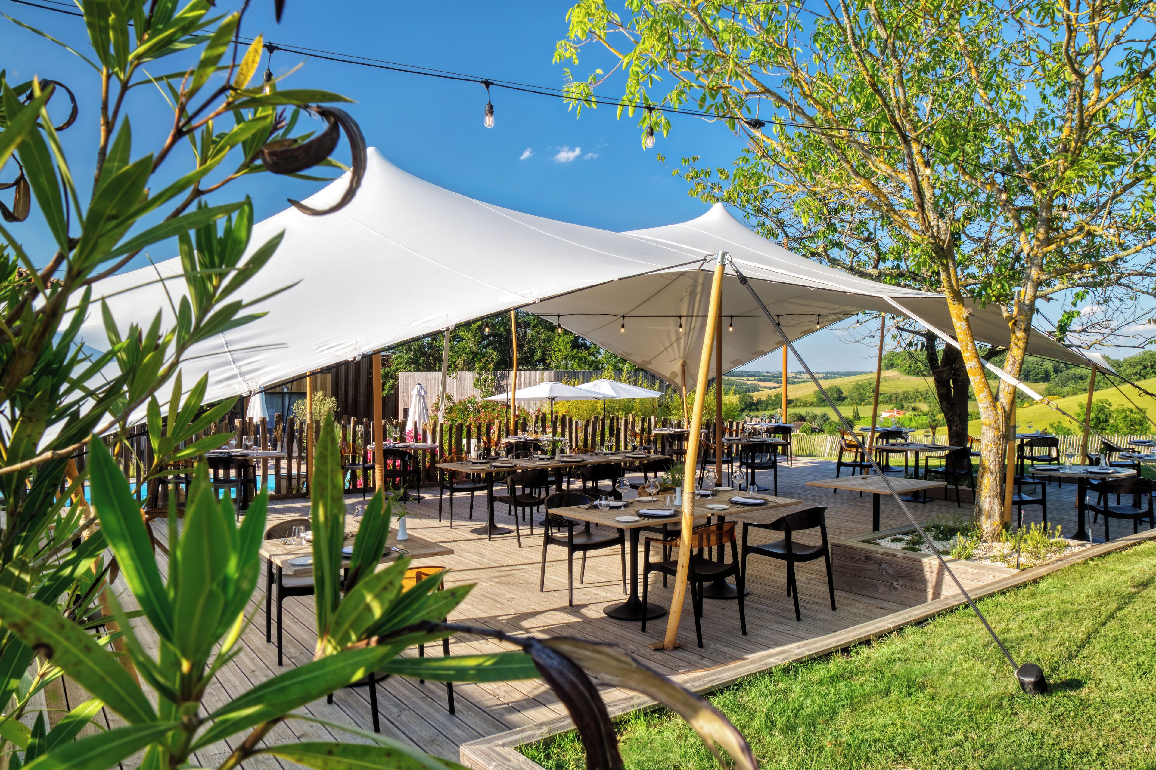 Un área de comedor al aire libre con mesas y sillas bajo grandes toldos blancos, rodeada de vegetación y árboles.