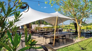 Un área de comedor al aire libre con mesas y sillas bajo grandes toldos blancos, rodeada de vegetación y árboles.