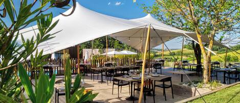 An outdoor dining area with tables and chairs under large white canopies, surrounded by greenery and trees.