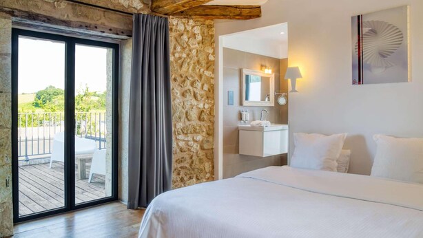 A bedroom with a large bed, a stone wall, a balcony with a table and chairs, and a bathroom with a mirror and sink. - Hotel Le Domaine De Baulieu (Auch)