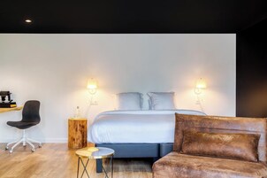 Una habitación de hotel moderna con una cama, dos lámparas de noche, una mesita de noche de madera, una mesa redonda de centro y un sillón de oficina gris, además de un sofá marrón.