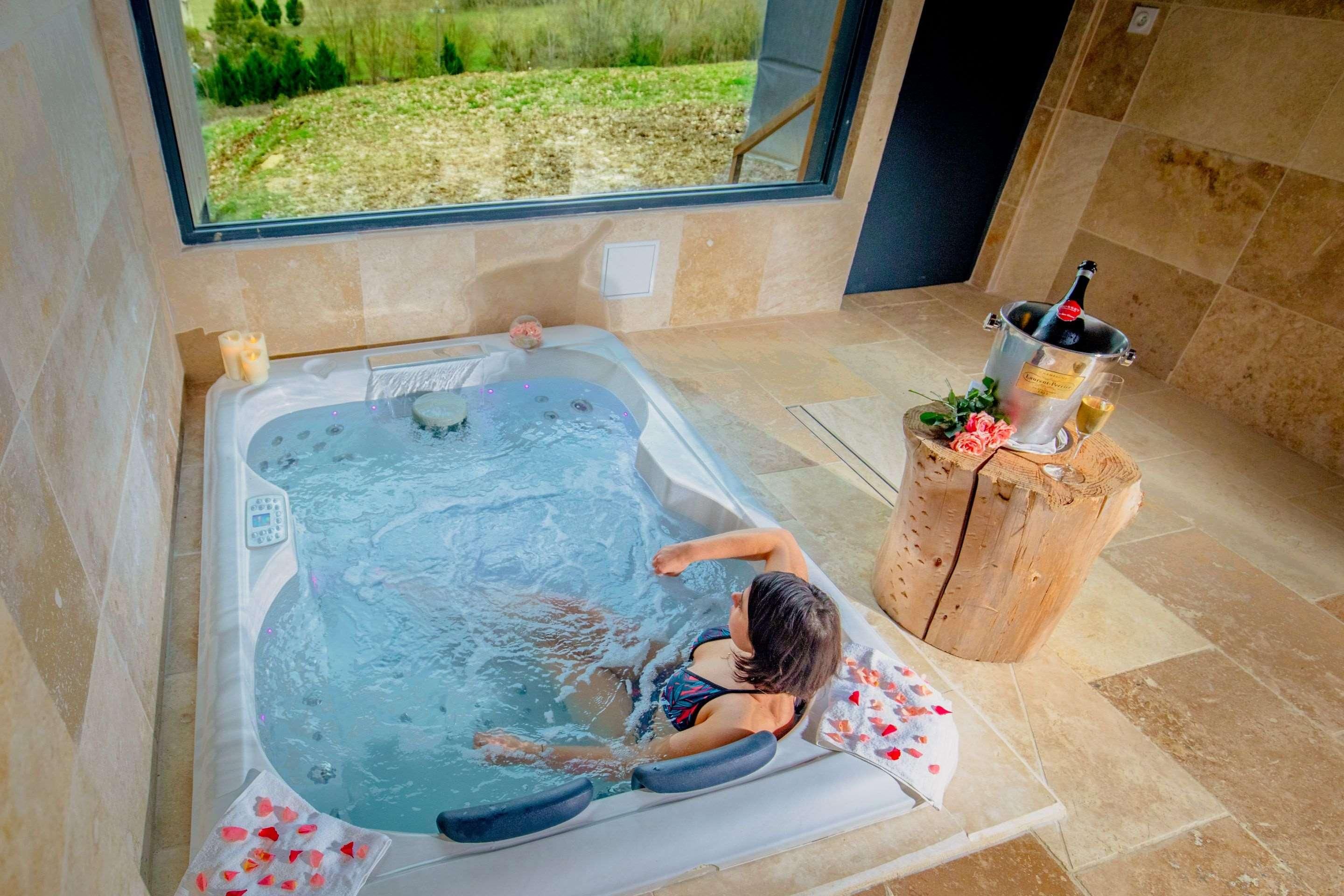 Una persona relajándose en un jacuzzi con vista a un jardín, un taburete de madera con una botella y un vaso de bebida.