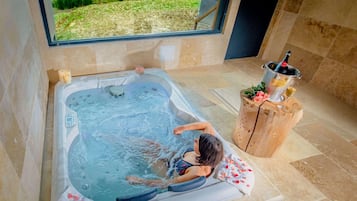 Una persona relajándose en un jacuzzi con vista a un jardín, un taburete de madera con una botella y un vaso de bebida.