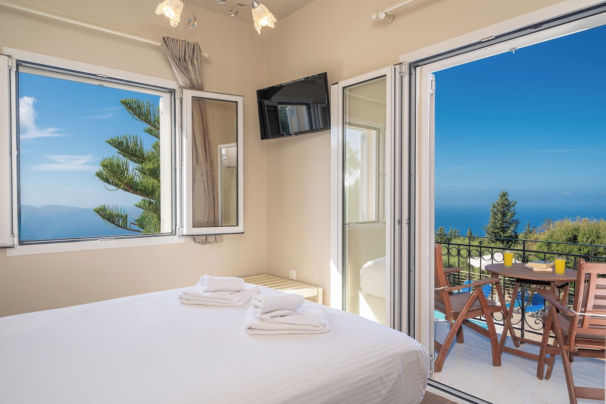 A bedroom with a large bed, a small table, and a chair. There is a balcony with a view of the sea and trees.