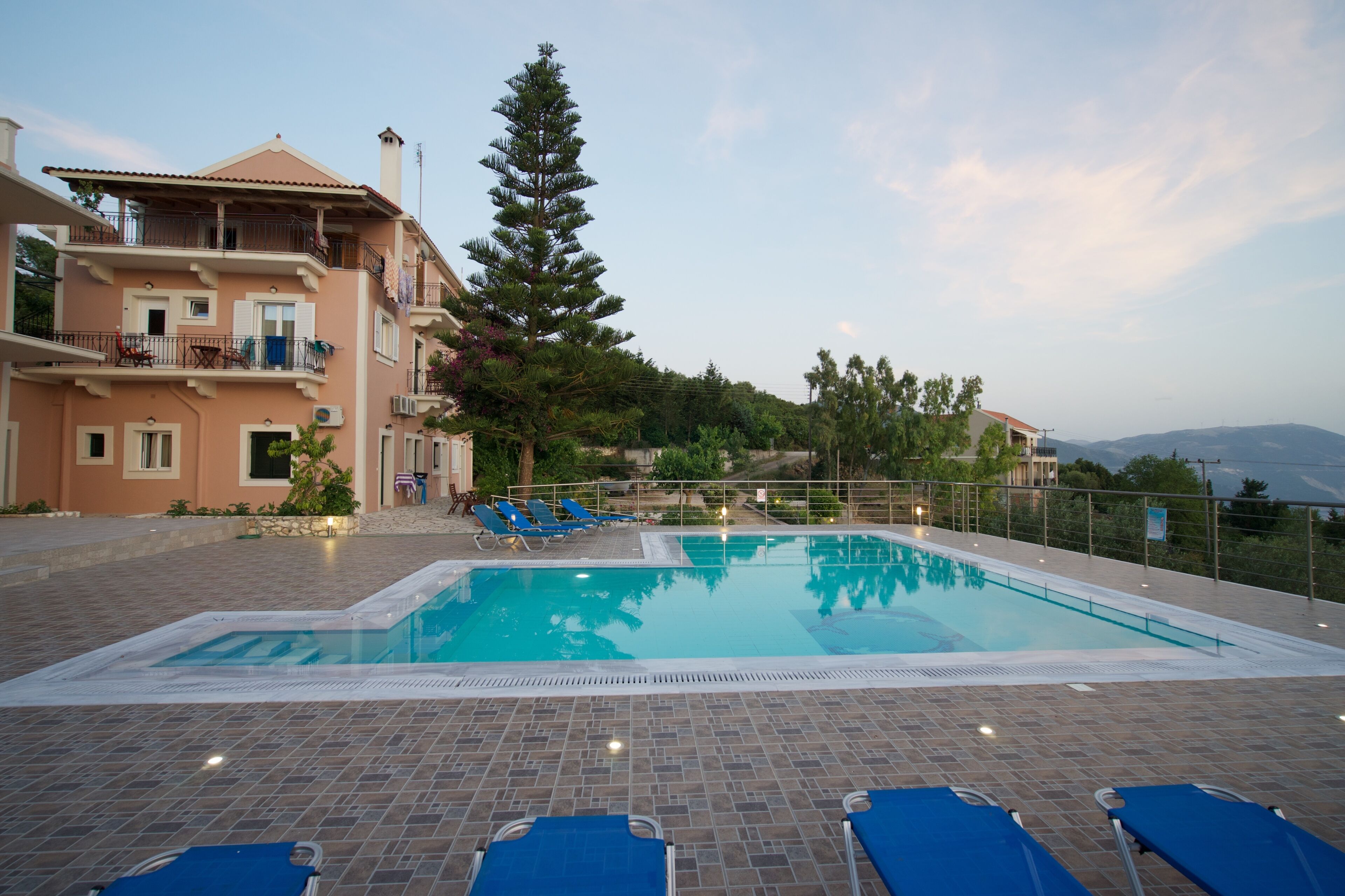 A swimming pool with blue lounge chairs, a large tree, and a building with balconies.
