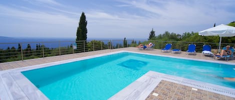 A rectangular swimming pool with a tiled deck, surrounded by blue lounge chairs and a white umbrella.
