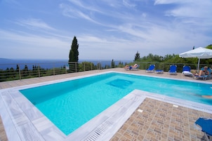 A rectangular swimming pool with a tiled deck, surrounded by blue lounge chairs and a white umbrella.