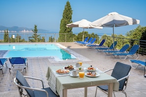 A poolside dining area with a table set for breakfast, blue lounge chairs, and umbrellas. - Studios Vrionis Panoramic Sunset (Kefalonia)