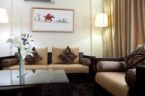 A hotel room with a sofa, armchairs, a glass coffee table, a vase with flowers, a framed picture on the wall, and a lamp. - Oak Plaza Hotel East Airport (Accra)
