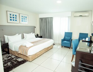 A hotel room with a large bed, a desk, two chairs, a window with curtains, and an air conditioning unit. - Oak Plaza Hotel East Airport (Accra)