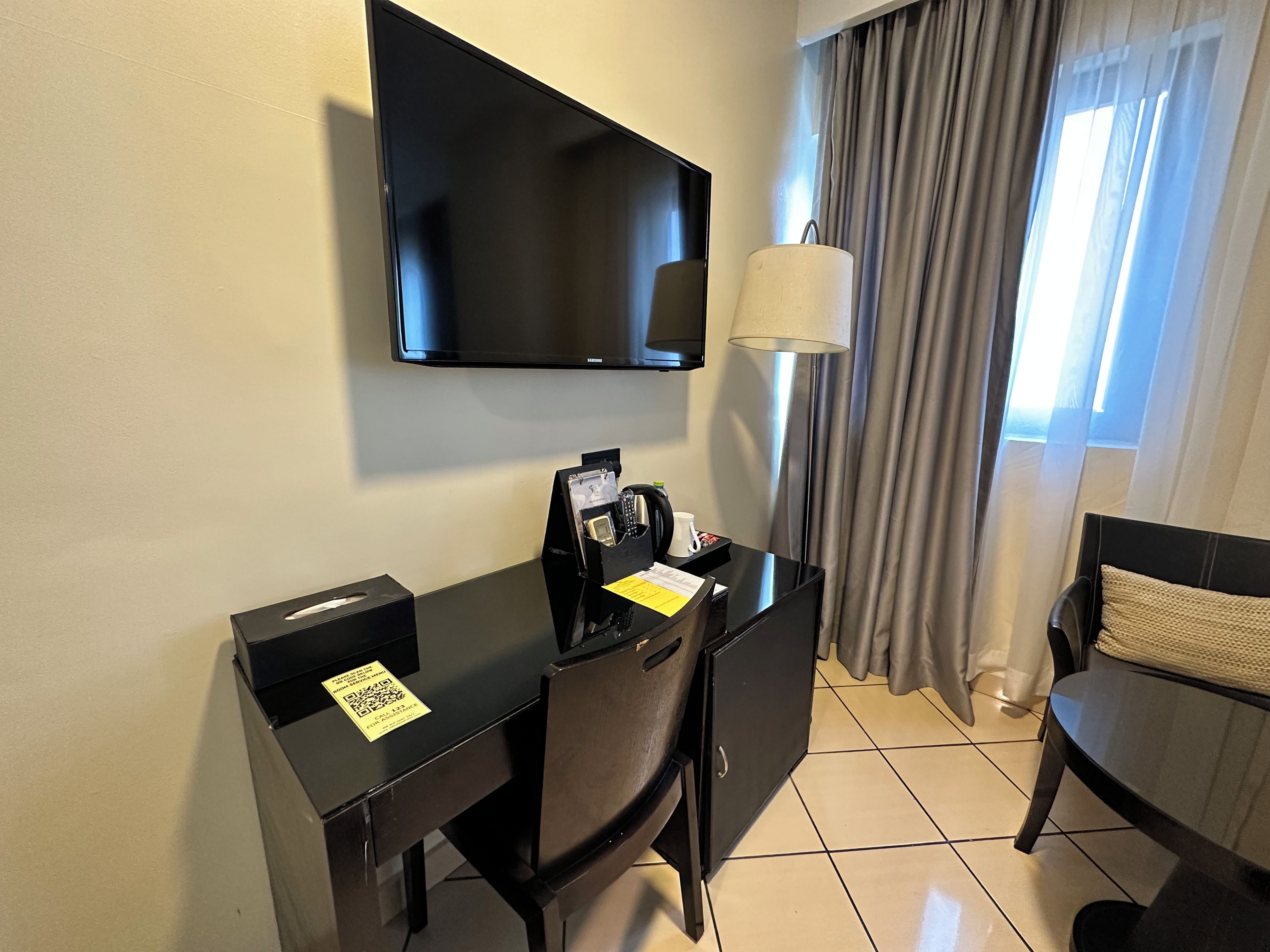 Habitación de hotel con una televisión de pantalla plana montada en la pared, un escritorio con teléfono y bloc de notas, una silla y una ventana con cortinas.