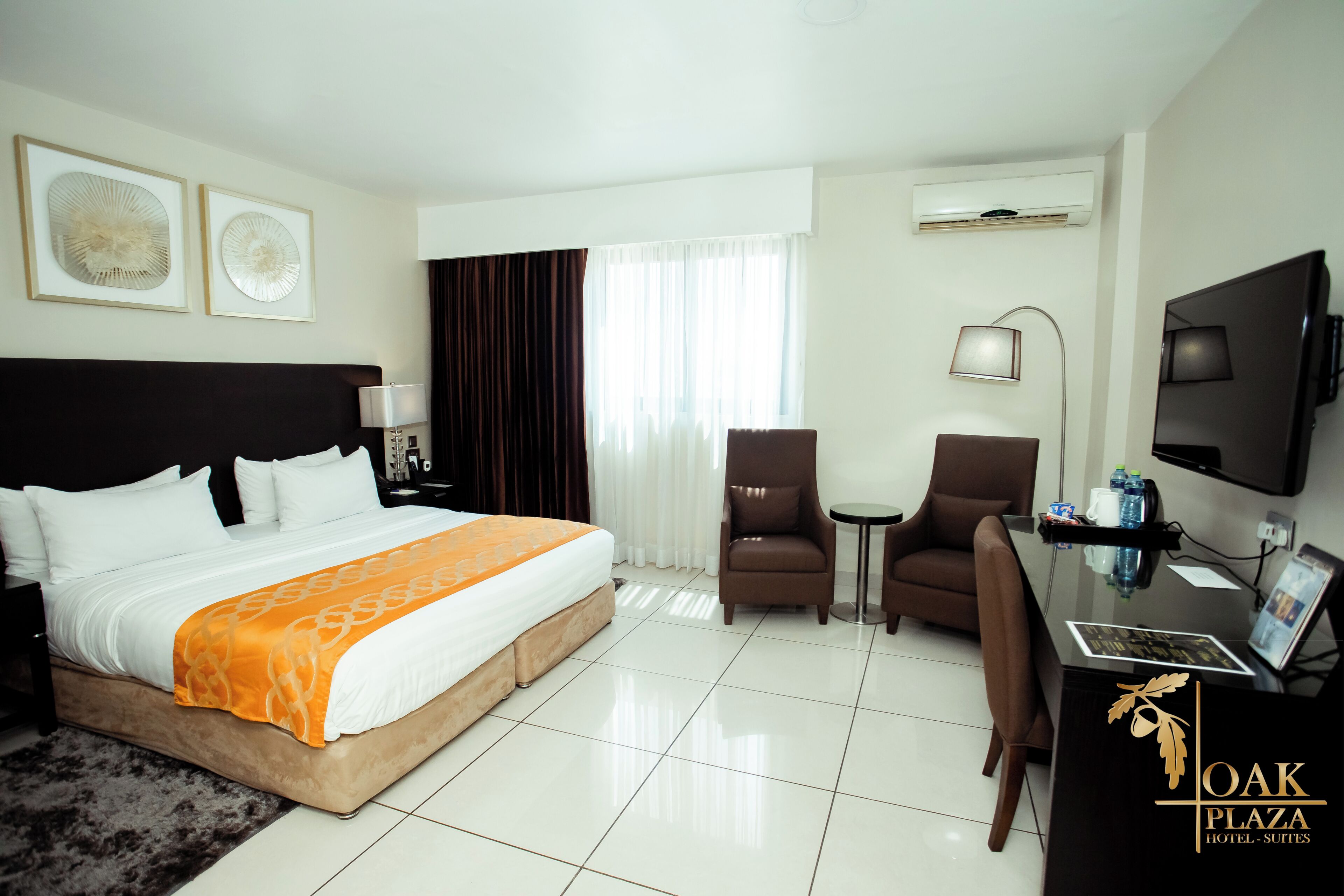 A hotel room with a bed, two chairs, a desk, a TV, and artwork on the wall.