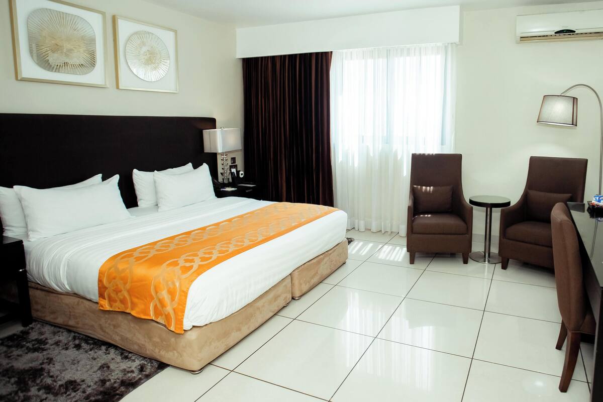 A hotel room with a bed, two chairs, a desk, a TV, and artwork on the wall.