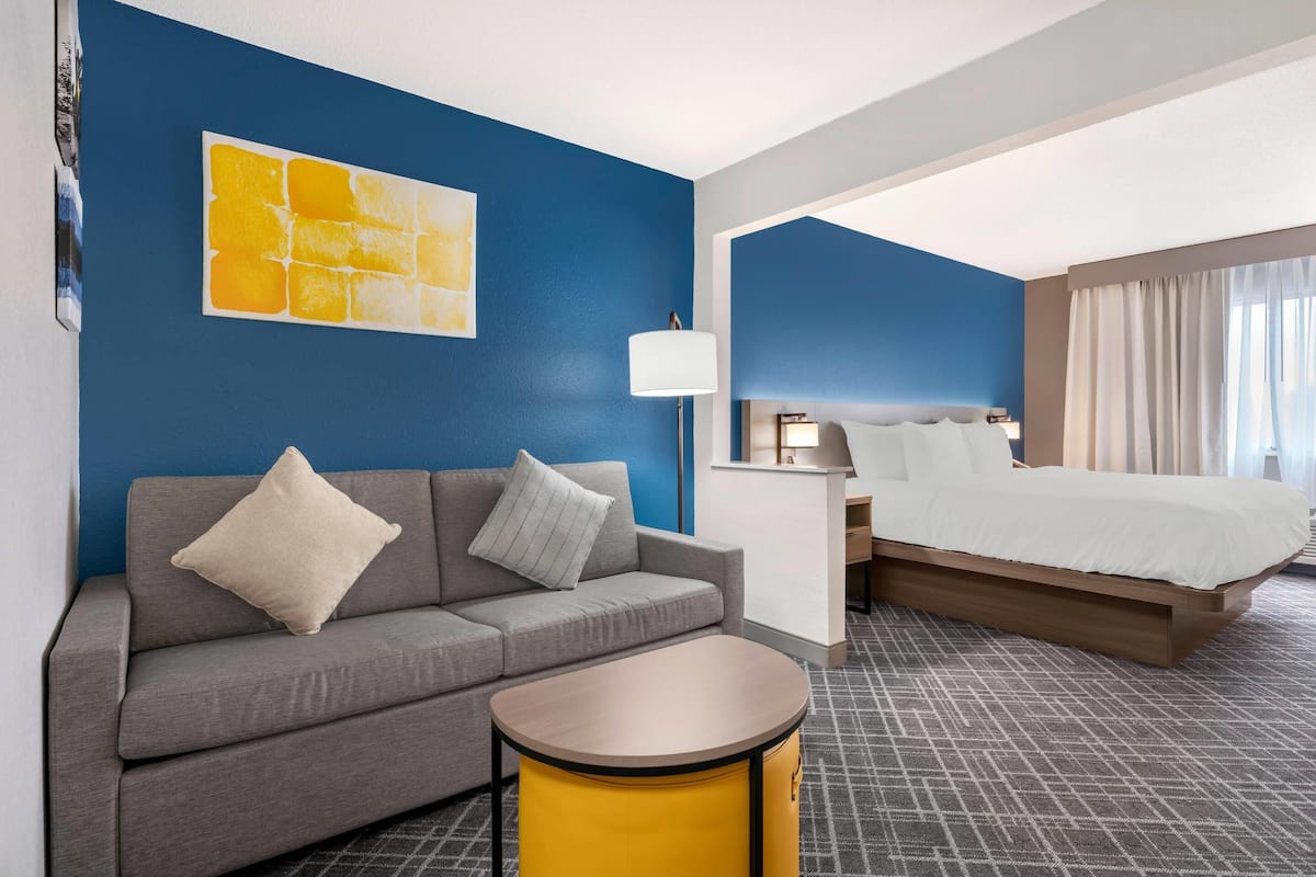 Quarto de hotel com um sofá cinza, uma mesa lateral amarela, uma cama com roupa de cama branca, uma mesinha de cabeceira, uma luminária e uma parede azul com uma obra de arte amarela.
