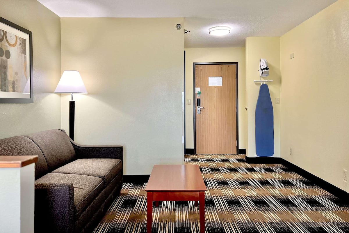 Quarto de hotel com sofá, uma mesa pequena, luminária, porta e um tapete estampado.
