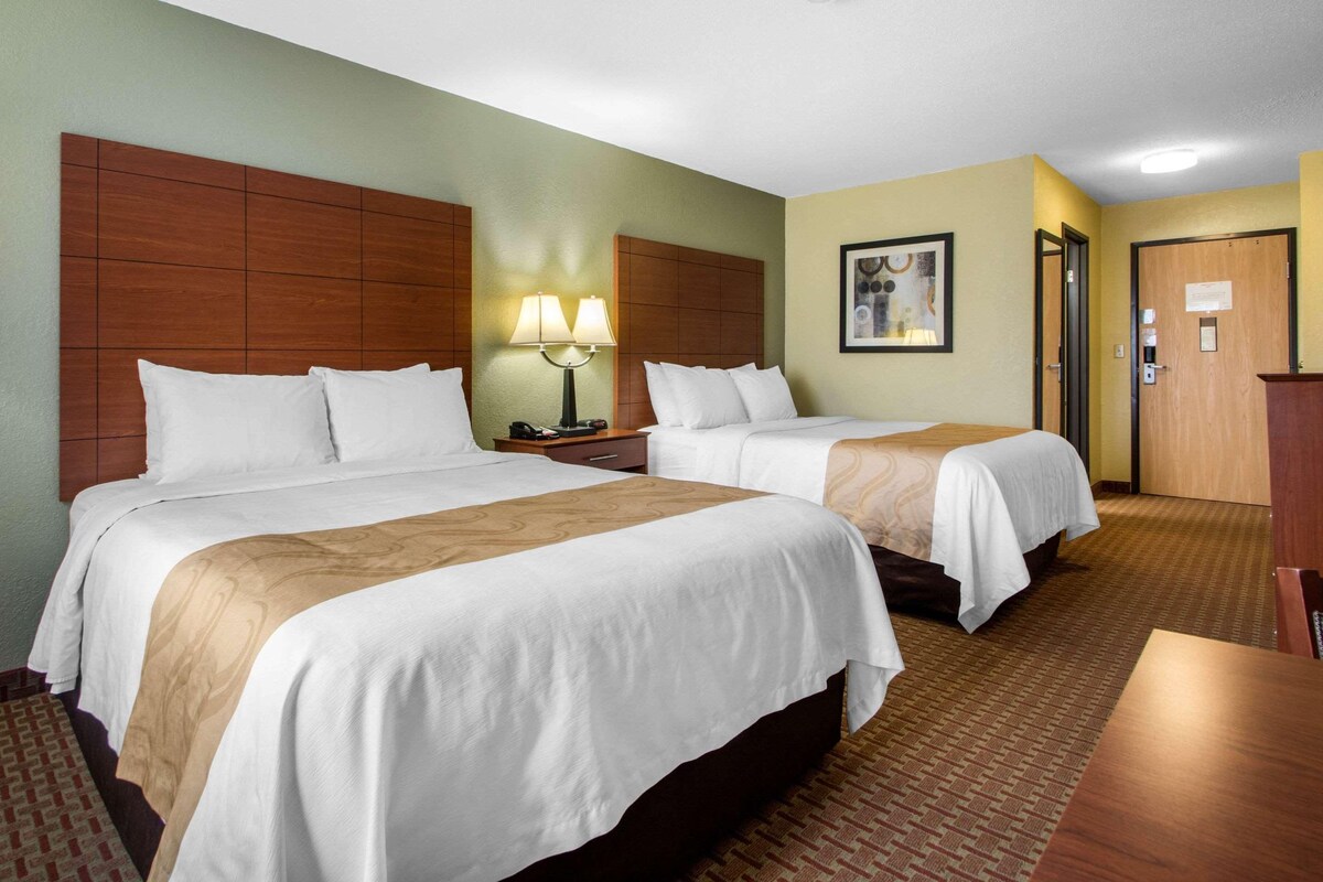 A hotel room with two beds, a nightstand with a lamp, a wooden headboard, a framed picture on the wall, and a door with a sign.